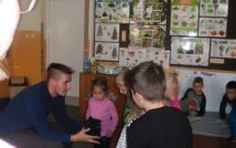 po lewej stronie zdjęcia siedzi listonosz i pokazuje jeden ze sprzętów do zbierania podpisów dotykowo. wokół niego stoją zaciekawione dzieci. w tle wielka tablica a na niej materiały dydaktyczne z przyrody.
