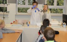 zdjęcie przedstawia klasę chemii w Liceum Ogólnokształcącym im. Mikołaja Kopernika. doł zdjęcia przedstawia siedzące tyłem dzieci. w centralnej górnej części zdjęcia stoją 2 uczennice i wykonują eksperyment wykorzystując ciecz i probówkę