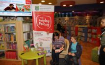 górny prawy róg przedstawia plakat z informacjami spotkania autorskiego wyświetlony na ekranie.lewy dół zdjęcia przedstawia niski stolik na którym znajdują się książki pisarki.centralna część zdjęcia to baner z projektu:Mali Wielcy Polacy. po prawej stronie siedzi pisarka, która podpisuje swoją książkę.koło niej siedzi fan.w tle oodział dla dzieci.