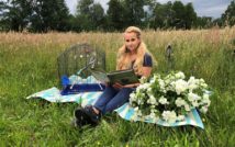 siedząca kobieta na kocu w trzyma książkę. koło niej ptaszek w klatce. za nią łąka