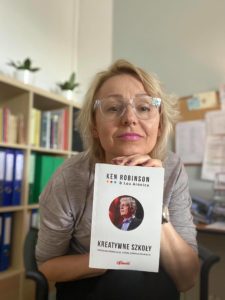 kobieta trzymająca książkę Kena Robinsona Kreatywne szkoły. W tle regał z ksiażkami