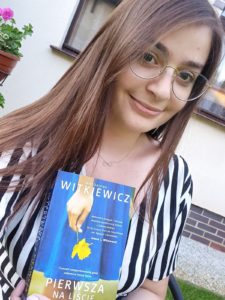 młoda dziewczyna z długimi włosami w czarnobiałej sukience, trzymająca w ręku książkę Magdaleny Witkiewicz Pierwsza w kolejce