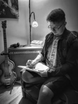 Młody mężczyzna w okularach siedzący w fotelu. W ręku trzyma kartki i ołówek. Z tyłu gitara i wzmacniacz. Na nim słuchawki, dwie książki. Za nim lampa. U góry fragment obrazu.