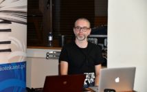Mężczyzna siedzi. Przed nim dwa laptopy. Za nim po lewej stronie fragment roll-upa.