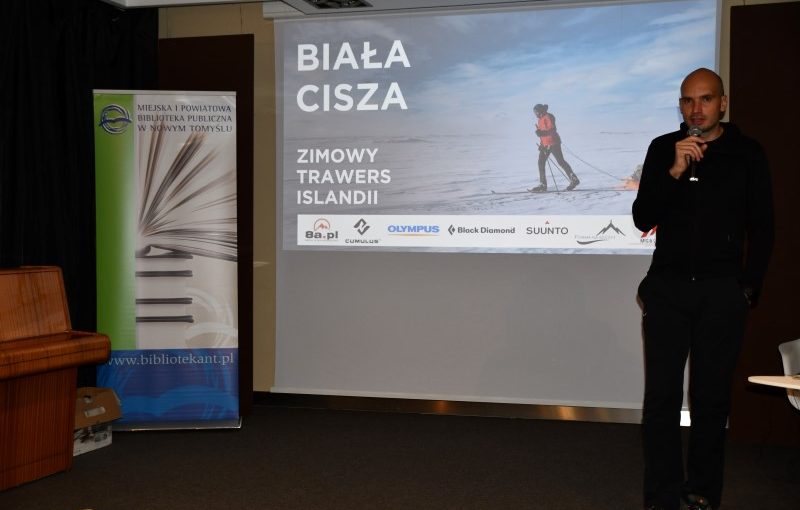Na wprost ekran. Na ekranie meżczyzna na nartach ciągnący sanie. Śnieg i niebo z chmurami. W górnym lewym rogu napis: Biała cisza. Niżej napis: Zimowy trawers Islandii. Na samym dole ekranu loga: 8a.pl, Eumulus, Olympus, Black Diamond, SUUNTO. Po prawej stronie zdjęcia człowiek z mikrofonem, po lewej roll-up. U góry roll-upu logo i napis: Miejska i Powiatowa Biblioteka Publiczna w Nowym Tomyślu. Na środku cztery leżące książki, na nich jedna otwarta. Na dole roll-upu napis: www.bibliotekant.pl. Po lewej stronie od roll-upu fragment pianina.