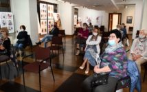 11 osób siedzących na sali w maseczkach na twarzach. Po lewej stronie i w głębi obrazki w antyramach.