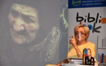 Siedząca kobieta z wzniesionym palcem do góry. Przed nią stolik, na stoliku książki i matrioszka z wizerunkiem mężczyzny. Z tyłu na ekranie starsza kobieta. Z tyłu po prawej stronie baner z napisem: bibliolook.