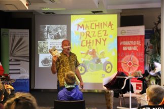 mężczyzna w koszuli z krótkim rękawem trzymający książkę. za nim na zdjęciu plakat ilustracyjny pary w motorze Machiną przez Chiny. w tle na zdjęciu