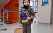 usmiechnięty chłopiec w czapce i kurtce w swojej lewej ręce trzyma dyplom.w prawej torbę z nagrodami. w tle hol biblioteki.
