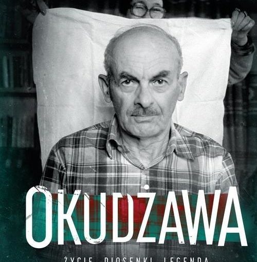 okładka książki, centralnie postać mężczyzny w kraciastej koszuli, za którym stojąca z tyłu, na tle regału z książkami kobieta, trzyma rozpoztartą białą tkaninę. U góry białe litery: Dmitrij Bykow , u dołu duży napis: Okudżawa i mniejsze litery: życie, piosenki, legenda