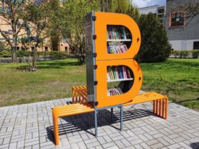 Zdjęcie bibliotecznego skweru, na króeym stoi półka bookcrossingowa w kształcie litery B. Wokół placyku wyłożonym kostką polbruk widac zieleń miejską