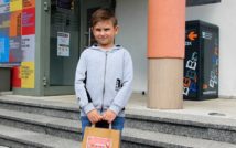 uśmiechnięty chłopiec.w obu rękach trzyma papierową torbę z nagrodą w środku.w tle front biblioteki