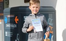 uśmiechnięty chłopiec.w lewej ręce trzyma papierową torbę z nagrodą w środku.w prawej trzyma dyplom za udział w konkursie.w tle wrzutnia biblioteczna.
