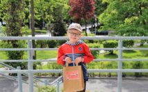 uśmiechnięty chłopiec w czapce i okularach. w obu rękach trzyma papierową torbę z nagrodą w środku.w tle zieleń miejska