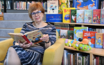 lewa strona zdjęcia: na fotelu siedzi kobieta w sukience i w okularach, trzyma otwartą książkę.W tle okładki książek.