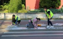 Trzy osoby w kamizelkach odblaskowych, przed przejściem dla pieszych na świeższym powietrzu. Dwie osoby klęczą i malują z tej pozycji po chodniku, jedna osoba stoi w pozycji pół zgiętej. W tle murek i zieleń.