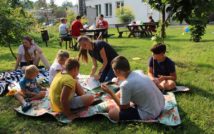 Zdjęcie przedstawia grupę osób siedzących na kocach, na trawie. W tle widoczne są inne osoby siedzące przy stolikach, budynek, krzewy i drzewa