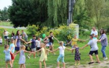 Zdjęcie przedstawia grupę osób - dzieci i dorosłych bawiących się w kółku i trzymających się za ręce. W tle widoczne inne osoby, stoły, drzewa, ulica