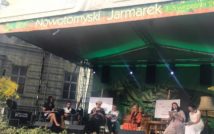 Siedem osób dorosłych siedzi na scenie z otwartymi książkami w rękach. U góry napis: Nowotomyski Jarmarek.
